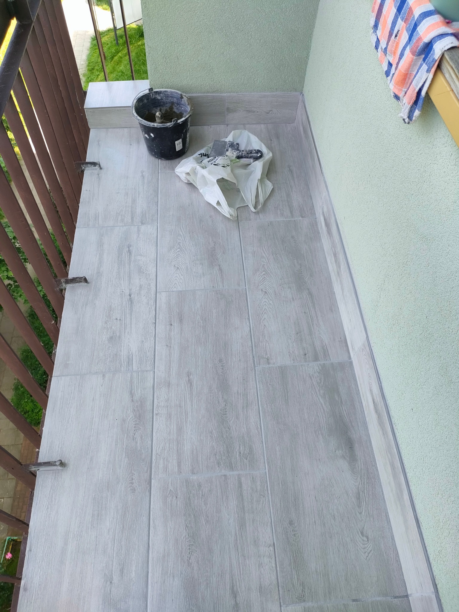 Ułożone szare płytki imitujące drewno na podłodze balkonu. Widoczne narzędzia i wiadro z zaprawą. Ściana pomalowana na jasnozielono.