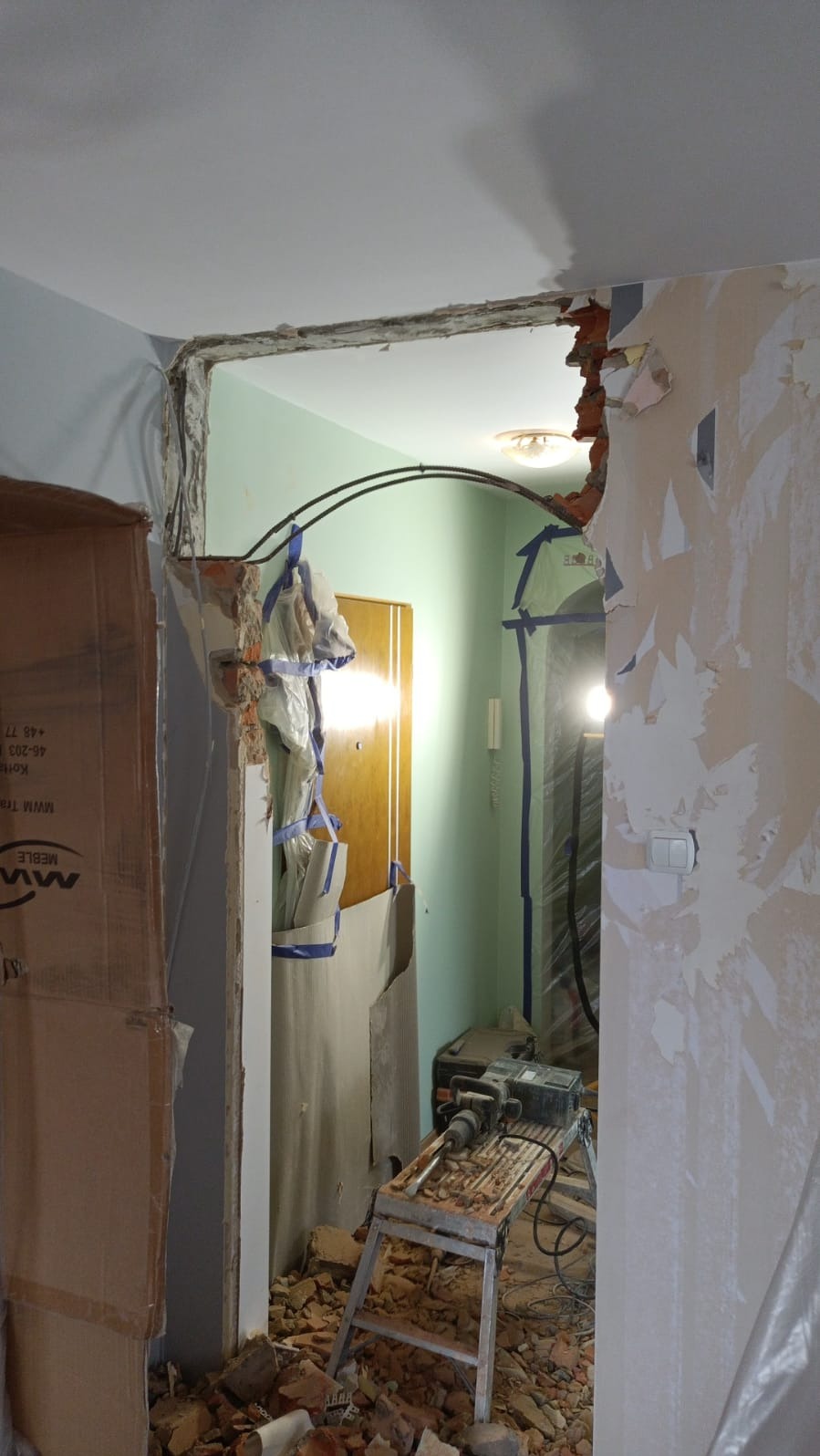 Demontaż futryny drzwiowej z widocznymi fragmentami muru, gruzem i narzędziami. Prace remontowe w trakcie, zabezpieczone folią i taśmą malarską.