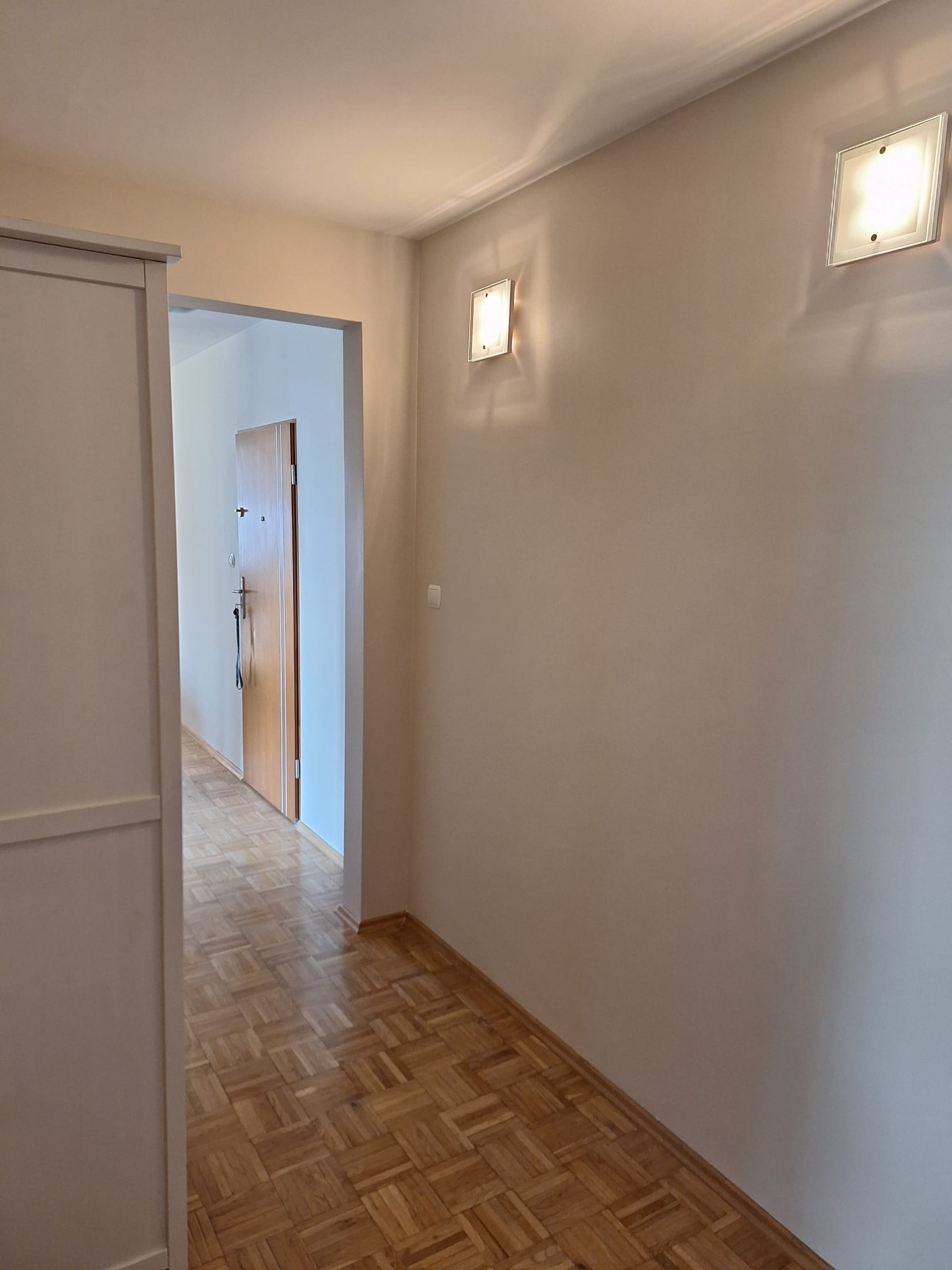 Jasne, świeżo pomalowane ściany w korytarzu z parkietem w Legnicy. Widoczne dwa kwadratowe kinkiety i drzwi w głębi. Minimalistyczny design.