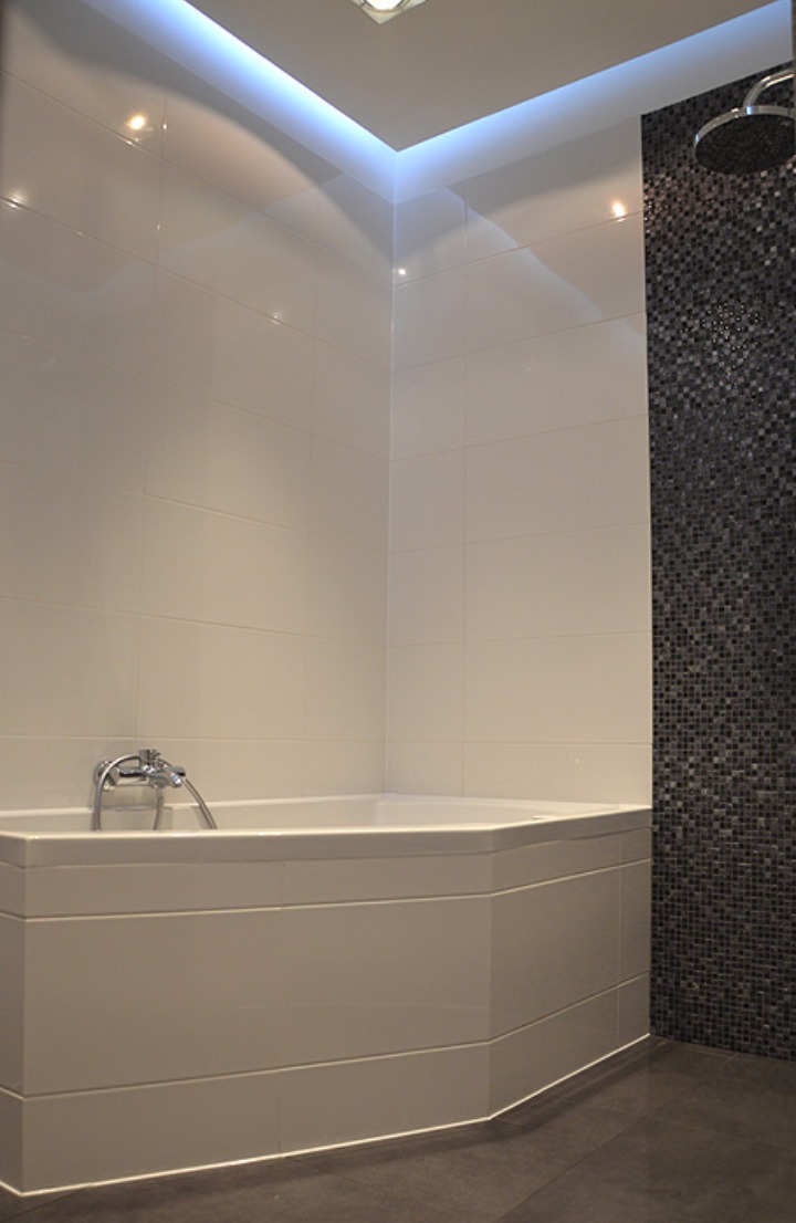 Nowoczesna łazienka z narożną wanną obudowaną panelami, białymi płytkami ściennymi i mozaiką w odcieniach szarości pod prysznicem, oświetlenie LED podsufitowe.
