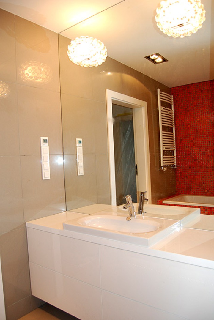Nowoczesna łazienka z dużym lustrem, białą szafką pod umywalką i czerwonymi płytkami mozaikowymi w strefie kąpieli, oświetlona kulistymi lampami sufitowymi.