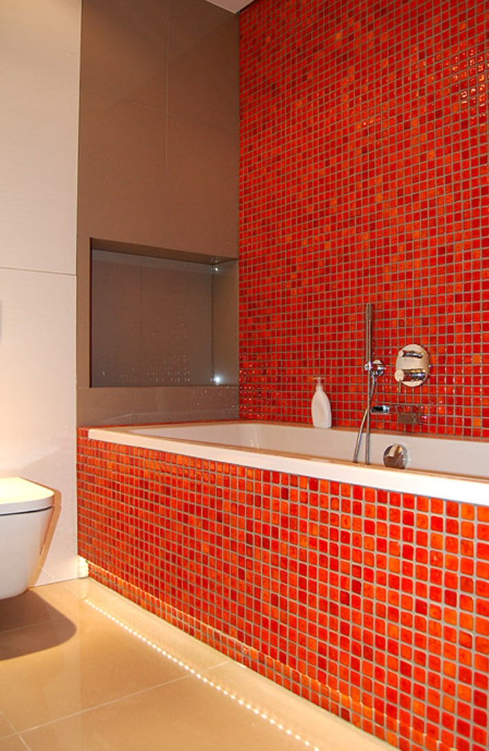 Nowoczesna łazienka z wanną obłożoną mozaiką w odcieniach czerwieni i pomarańczy, podświetlona listwą LED przy podłodze, z wnęką w ścianie wykończoną w szarym kolorze.