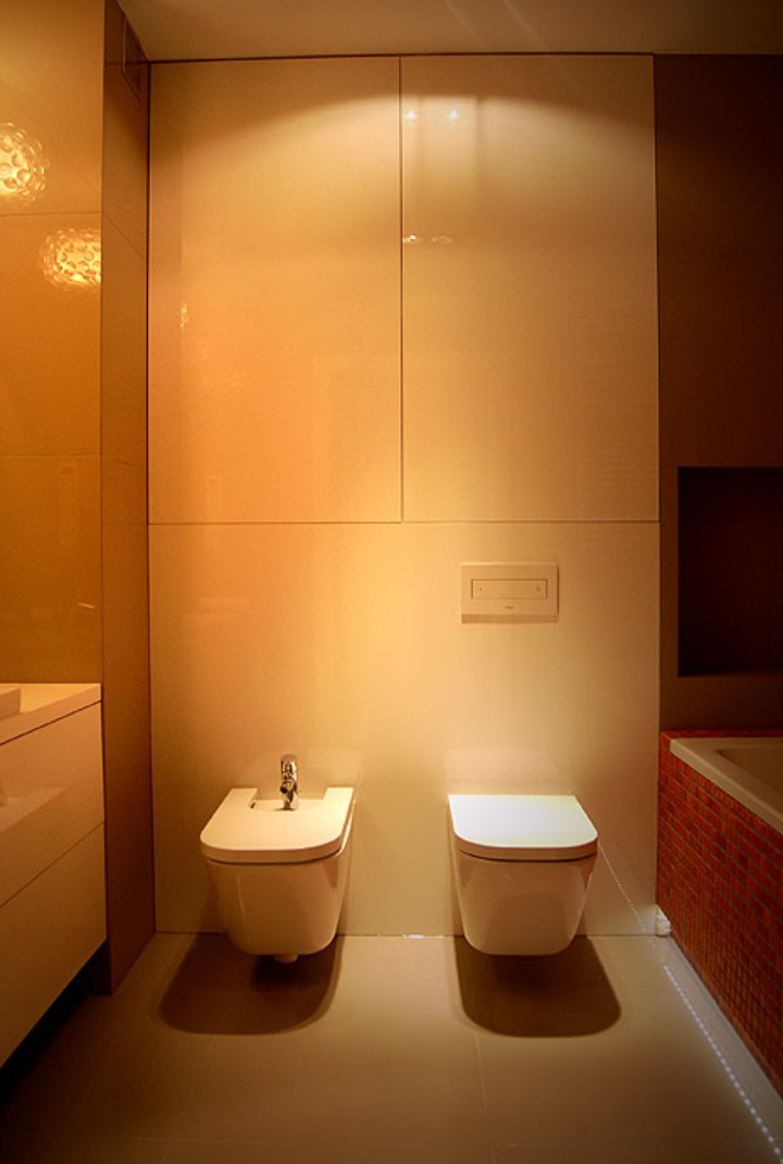 Nowoczesna łazienka z białymi, podwieszanymi toaletami i bidetem, oświetlona ciepłym światłem, z minimalistyczną zabudową meblową i czerwono-mozaikową obudową wanny.
