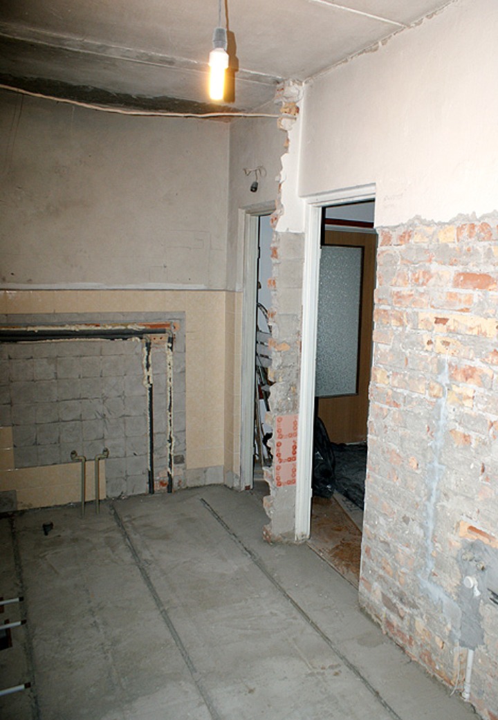 Wnętrze w trakcie demontażu, odsłonięta cegła, surowy beton na podłodze, widoczne instalacje, pojedyncza żarówka oświetla pomieszczenie, ślady po płytkach na ścianie.