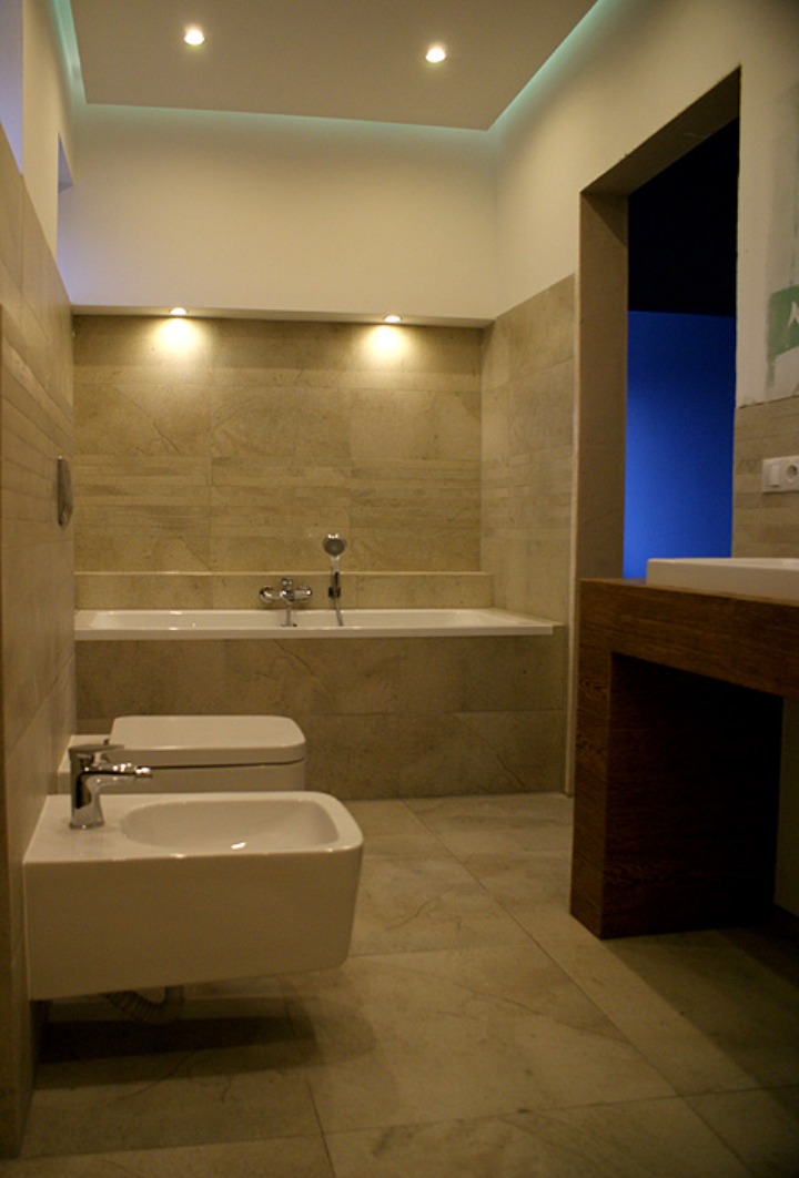 Nowoczesna łazienka z beżowymi płytkami, podwieszanym bidetem i toaletą, oraz drewnianą umywalką, oświetlona punktowymi lampami sufitowymi i listwą LED.