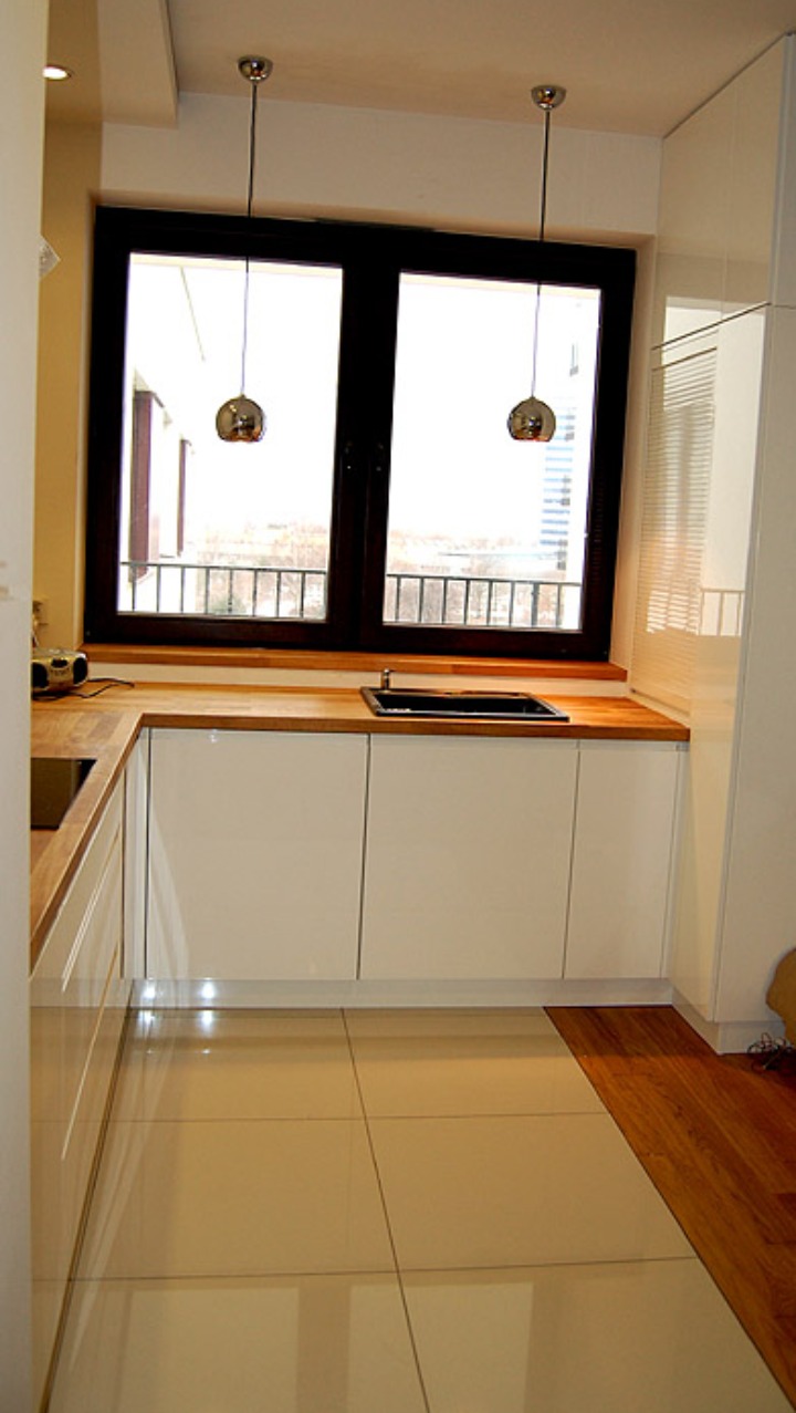 Nowoczesna kuchnia z białymi szafkami, drewnianym blatem i czarnym zlewem pod dużym oknem z ciemną ramą, oświetlona dwoma wiszącymi lampami w stylu retro.