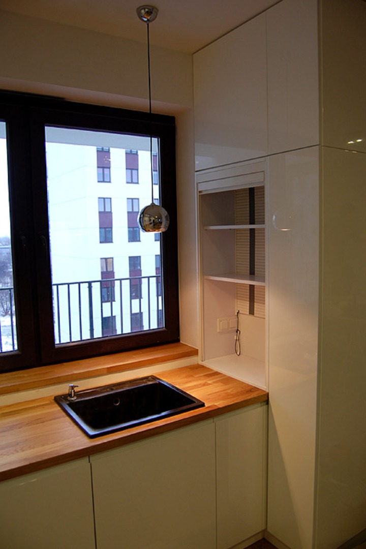 Nowoczesna kuchnia z czarnym zlewem pod oknem i białymi szafkami z połyskiem, drewniany blat, wisząca lampa w kształcie kuli.