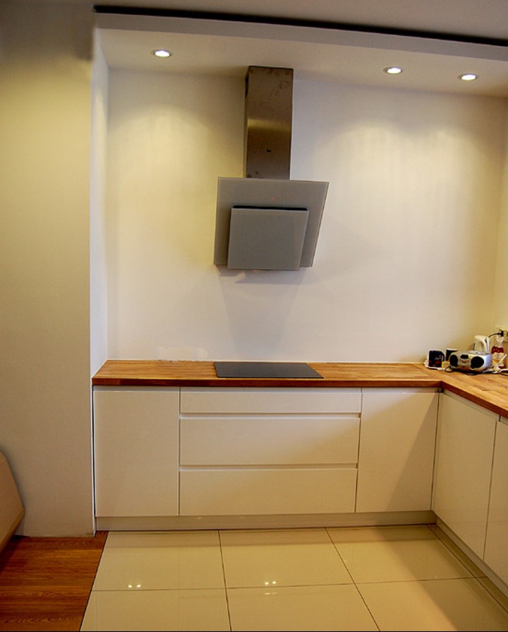 Nowoczesna kuchnia z białymi szafkami, drewnianym blatem i szklanym okapem, widok na płytę indukcyjną i fragment podłogi z płytek.