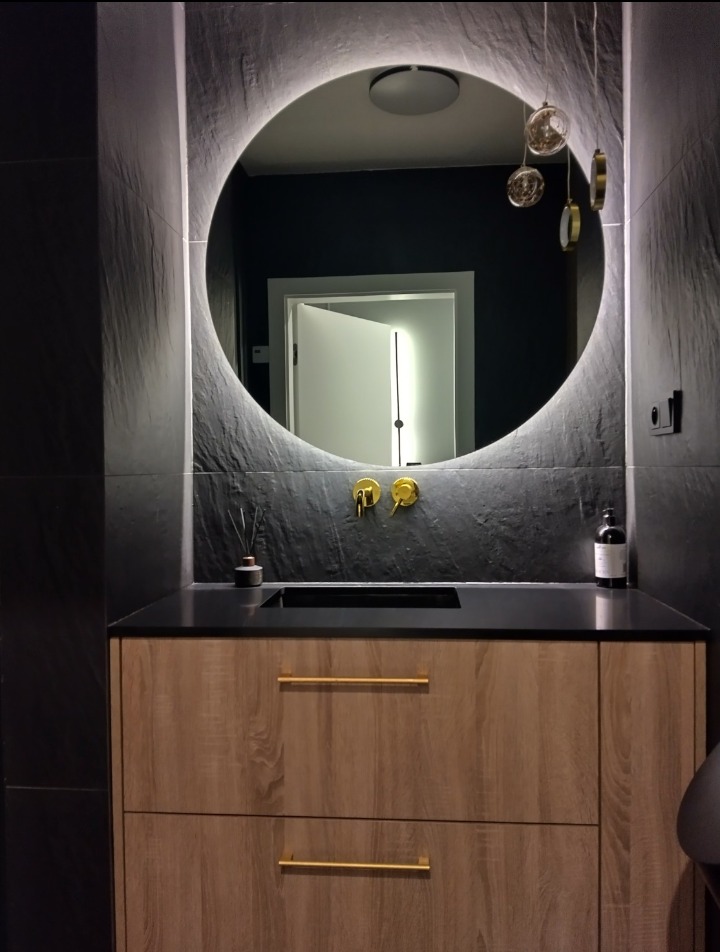 Nowoczesna łazienka z okrągłym lustrem podświetlanym LED, złotą armaturą i szafką z drewnopodobnym frontem, czarne płytki imitujące kamień na ścianach.