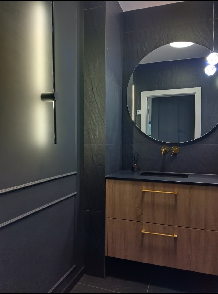 Nowoczesna łazienka z ciemnoszarymi płytkami imitującymi łupek, okrągłym lustrem z podświetleniem LED, drewnianą szafką pod umywalką z złotymi uchwytami i minimalistyczną lampą ścienną.