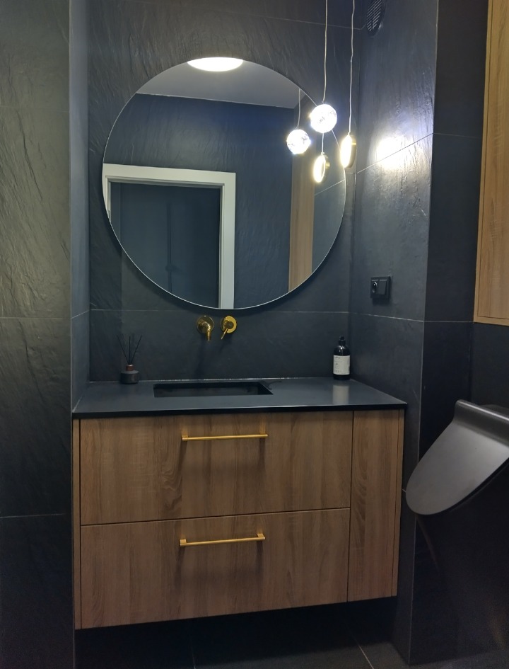 Nowoczesna łazienka w Kadzidle: Szafka pod umywalkę z drewnianymi frontami i złotymi uchwytami, okrągłe lustro z podświetleniem LED, czarne płytki na ścianach.