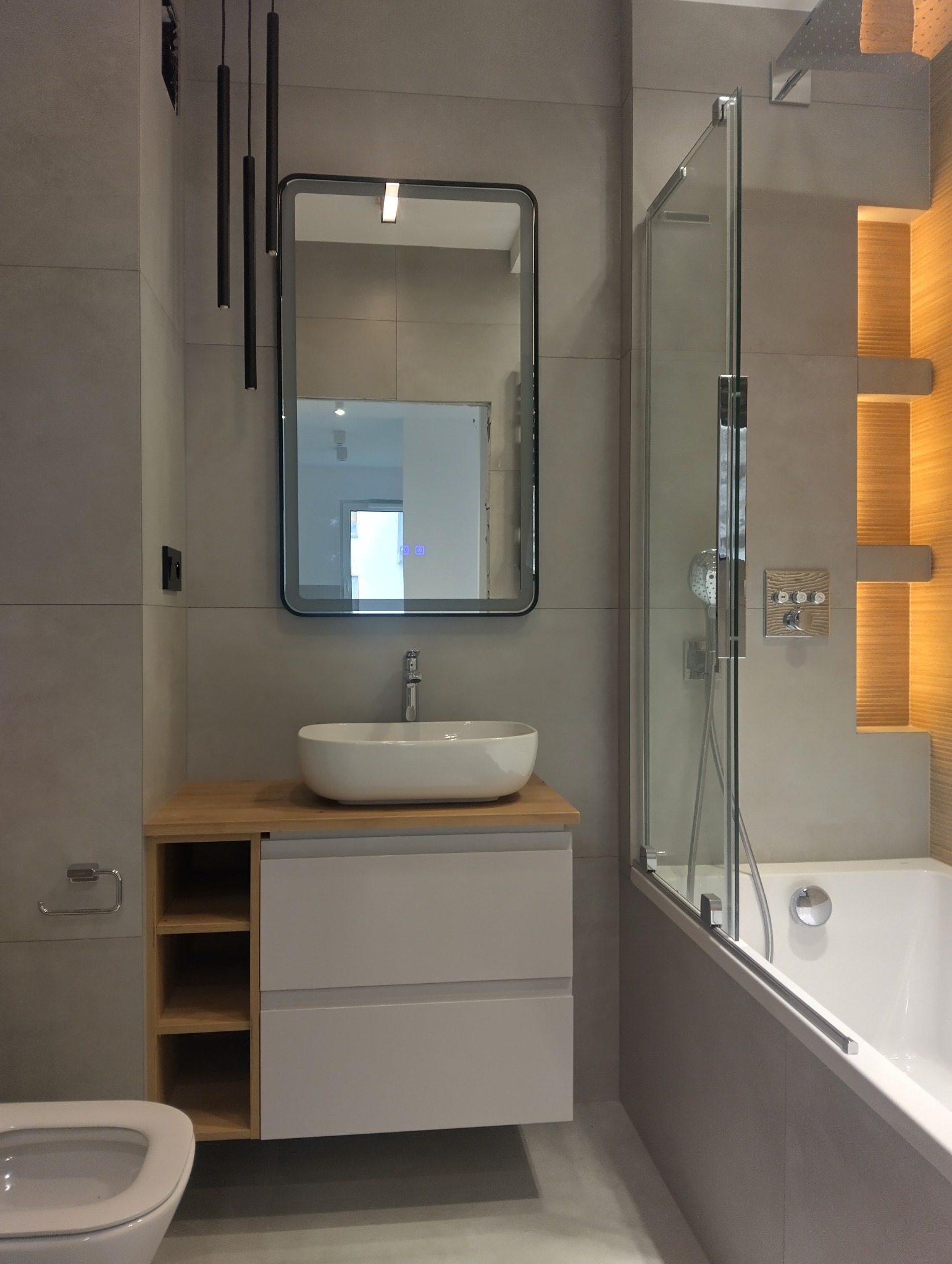Nowoczesna łazienka z szarą glazurą, owalną umywalką nablatową i kabiną prysznicową ze szklaną ścianką. Minimalistyczne oświetlenie i drewniane akcenty.