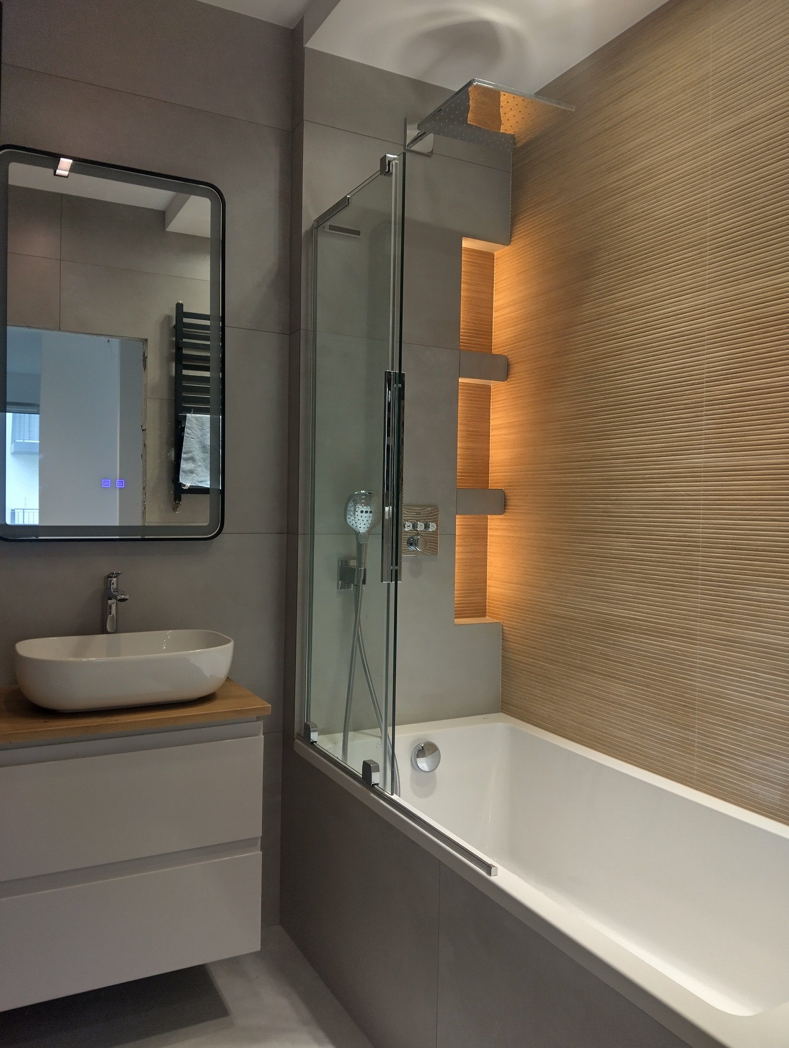 Nowoczesna łazienka z wanną i szklanym parawanem, umywalką na drewnianym blacie i podświetlanymi półkami. Szare płytki i beżowa ściana z poziomymi żłobieniami.