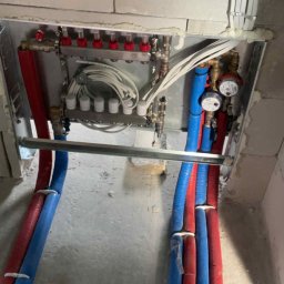 Hydro-Therm Mikołaj Janca - Rozdzielacz ogrzewania podłogowego z rurami w kolorze niebieskim i czerwonym, zamontowany w surowym pomieszczeniu. Widoczne białe przewody i uszczelnienia.