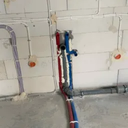 Instalacja wodna i elektryczna w surowym stanie: rury czerwone i niebieskie przymocowane do ściany z betonu komórkowego, przewody elektryczne w białych listwach.
