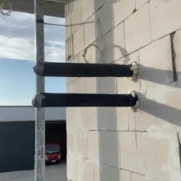 Dwie poziome rury, prawdopodobnie instalacyjne, przymocowane do ściany z betonu komórkowego, zabezpieczone pianką montażową. Na zewnątrz widoczny kontener i budynek.