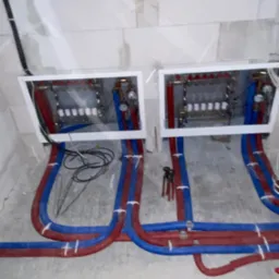 Rozdzielacze ogrzewania podłogowego w białych ramkach, z czerwonymi i niebieskimi rurami, na surowym betonie, przed wylaniem posadzki. Widoczne narzędzia.