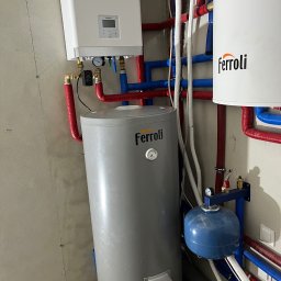 Hydro-Therm Mikołaj Janca - Nowoczesna instalacja grzewcza z piecem gazowym Kaisai i zasobnikami Ferroli, z widocznymi rurami w kolorach czerwonym i niebieskim, zamontowana na surowej ścianie.