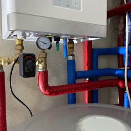Hydro-Therm Mikołaj Janca - Nowoczesny piec gazowy z manometrem i zabezpieczeniem Goshe, podłączony do instalacji z rur w izolacji czerwonej i niebieskiej. Widok z dołu, detale przyłączeń.
