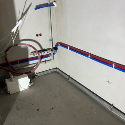 Hydro-Therm Mikołaj Janca - Szare rury kanalizacyjne i czerwono-niebieskie rury wodne zamontowane na ścianie w surowym pomieszczeniu z betonową podłogą. Widoczna pompa.