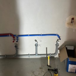 Hydro-Therm Mikołaj Janca - Szare i kolorowe rury instalacji wodnej zamocowane na ścianie, widoczne narzędzia pracy i karton. Instalacja wykonana w surowym pomieszczeniu.