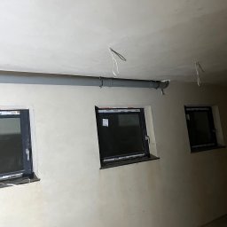 Hydro-Therm Mikołaj Janca - Szara rura instalacyjna zamontowana pod sufitem w pomieszczeniu z trzema oknami o czarnych ramach. Ściany w odcieniach beżu, sufit biały. Widoczne elementy mocujące rurę.