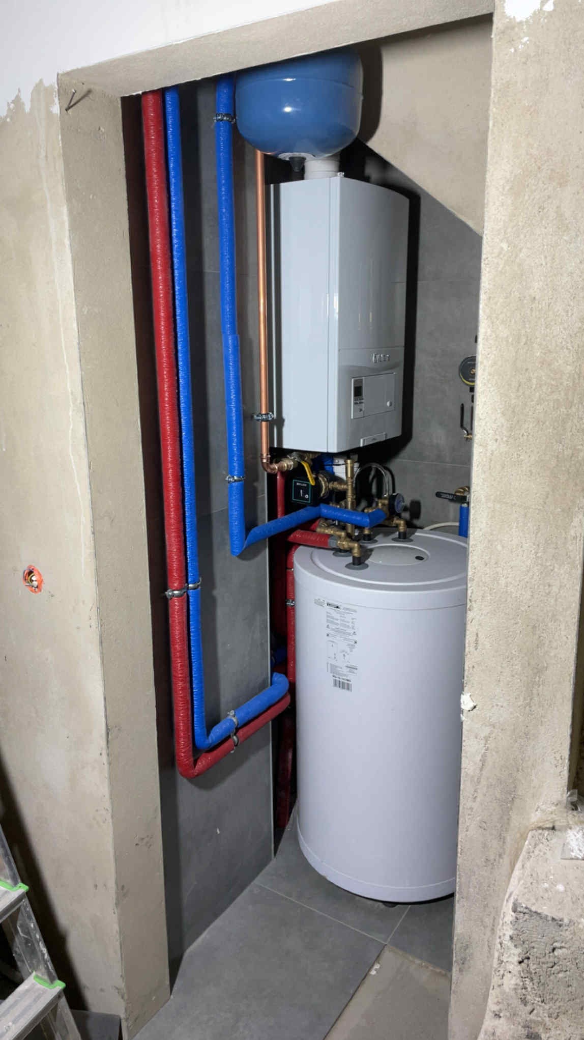 Nowoczesny piec gazowy i zasobnik ciepłej wody, z widocznymi rurami w kolorach niebieskim i czerwonym, zamontowane w pomieszczeniu z szarą glazurą.