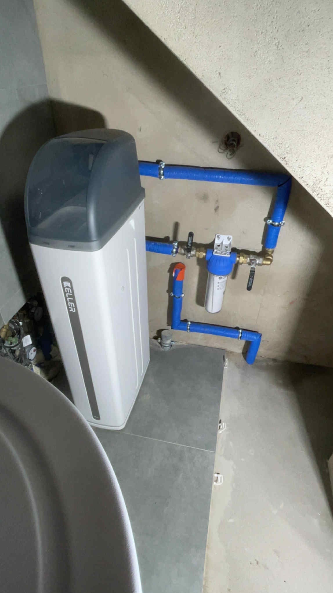 Nowoczesny zmiękczacz wody Keller z niebieskimi rurami instalacji wodnej, zamontowany w narożniku pomieszczenia. Filtr wody na ścianie.