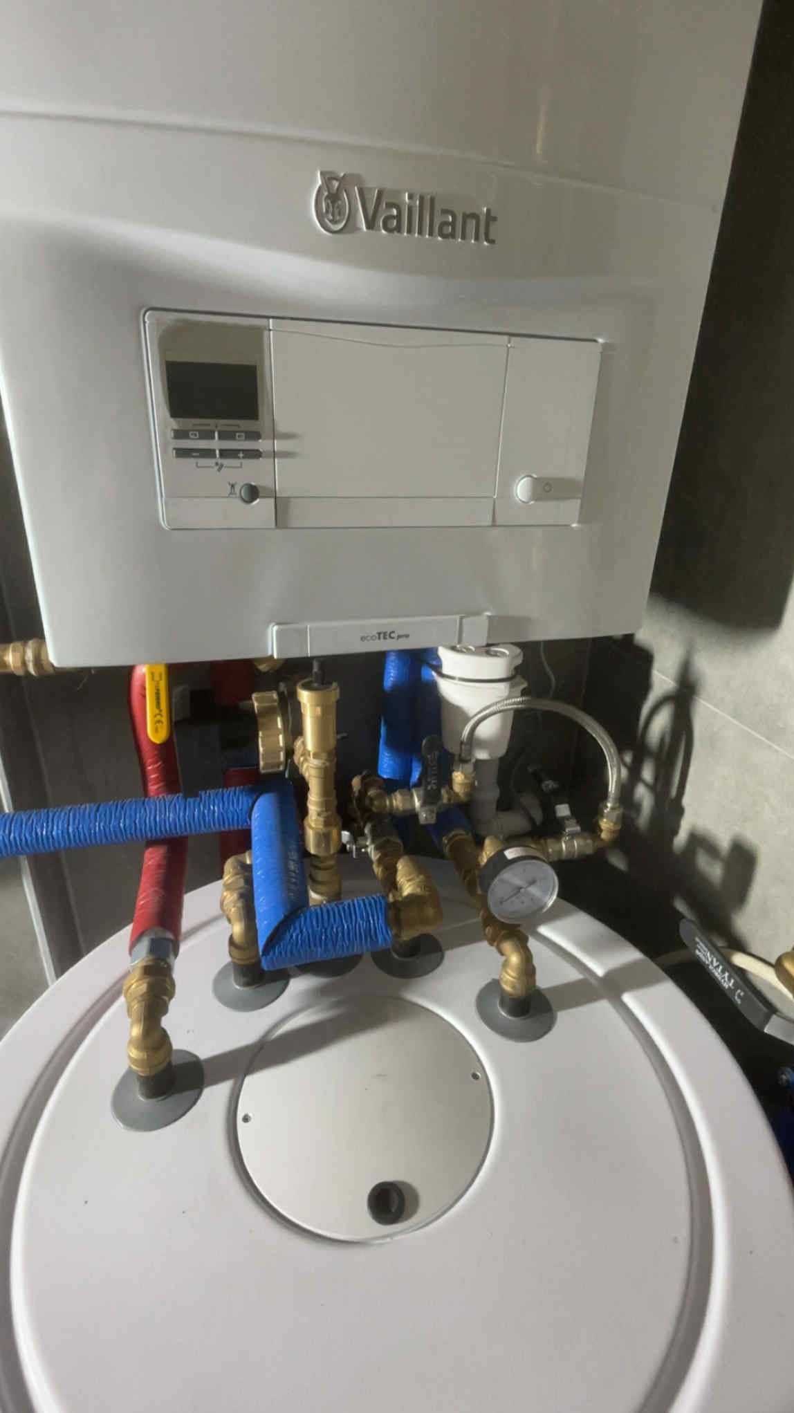 Biały piec gazowy Vaillant z podłączonymi rurami miedzianymi i niebieskimi, karbowanymi. Widoczne zawory i manometr. Całość na tle szarej ściany.