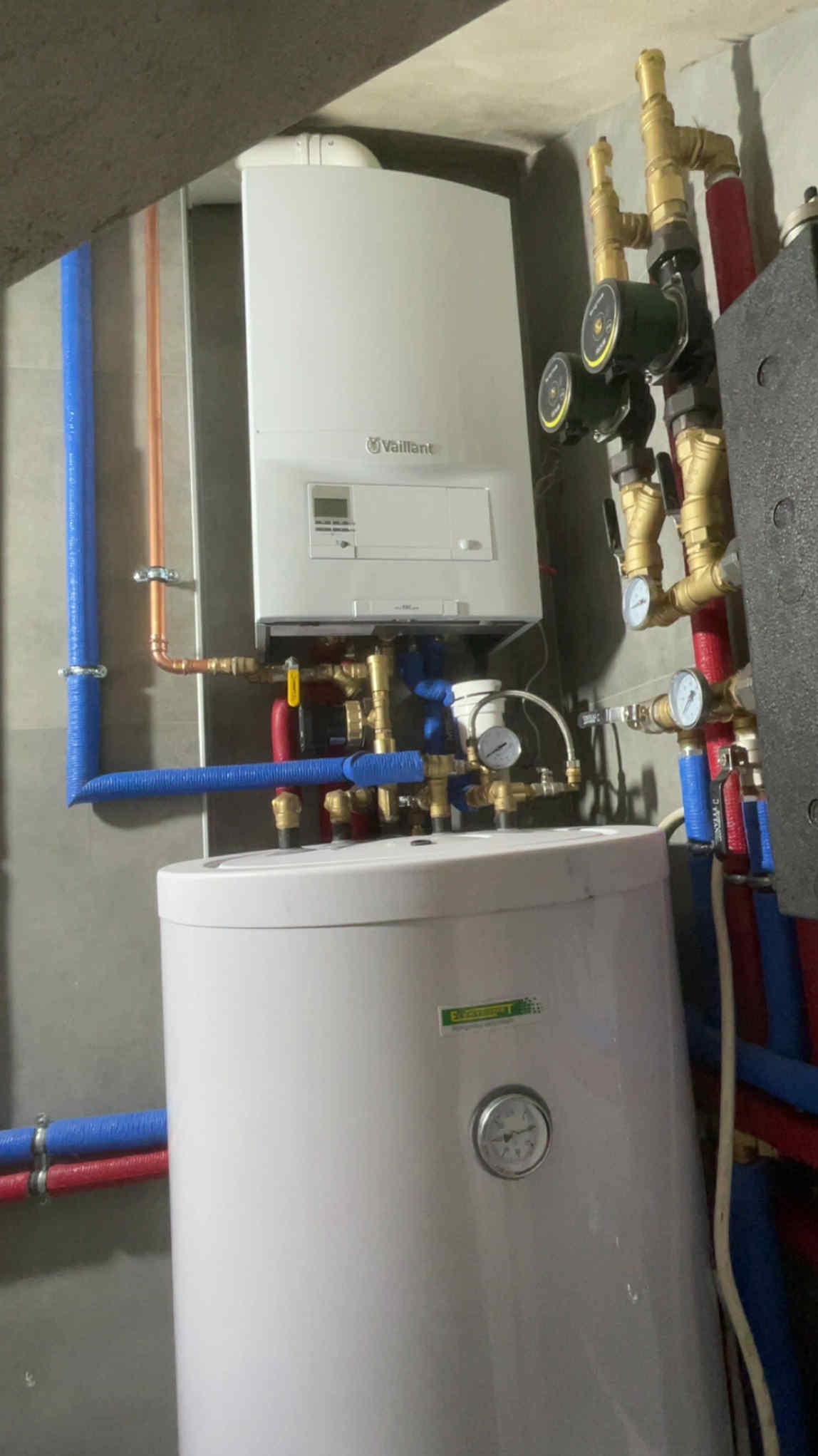 Nowoczesny piec gazowy Vaillant z podłączonym zasobnikiem ciepłej wody, instalacja z rur miedzianych i pex, manometry, pompy obiegowe, szare ściany w tle.