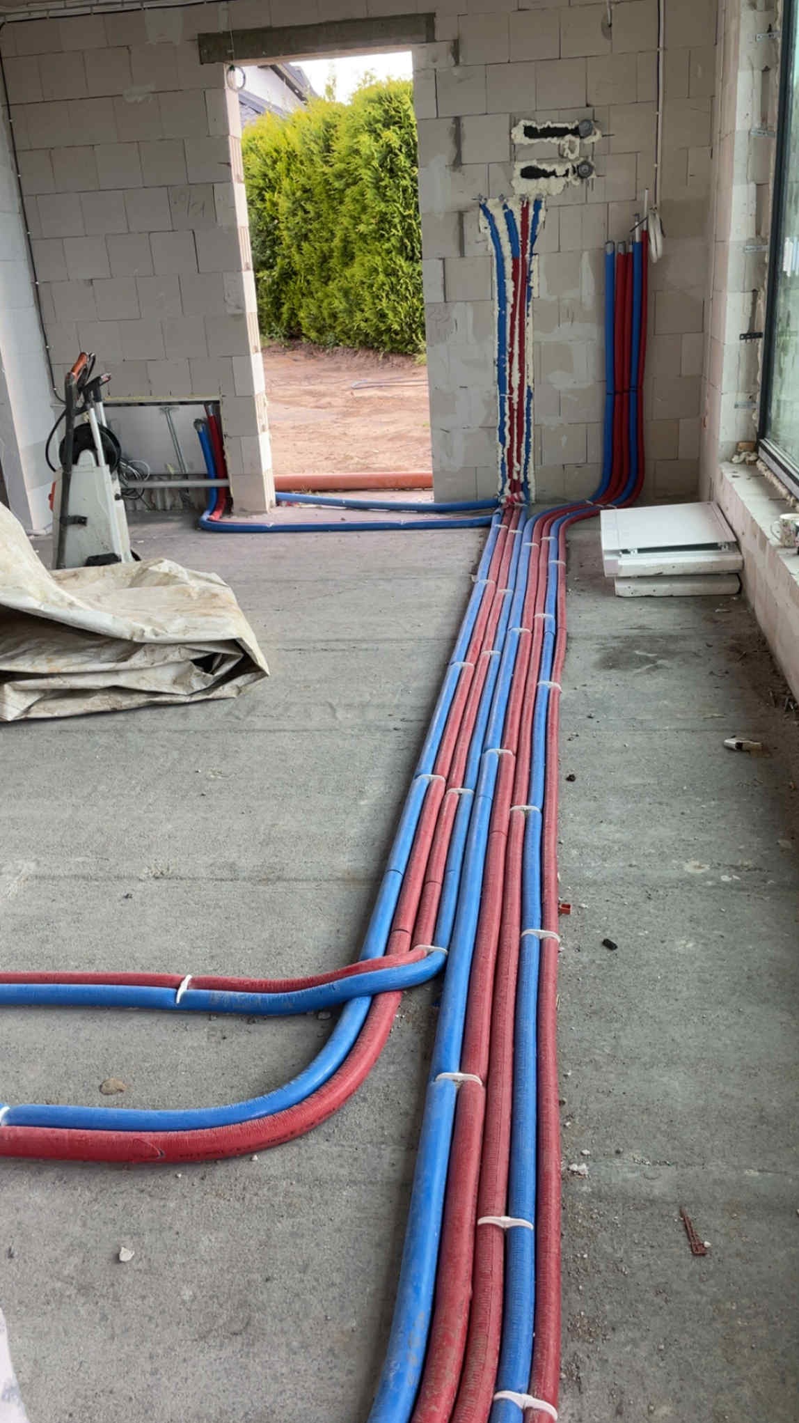 Układanie rur ogrzewania podłogowego w surowym wnętrzu. Czerwone i niebieskie rury prowadzone równolegle, mocowane białymi opaskami. Widoczne ściany z bloczków i okno.
