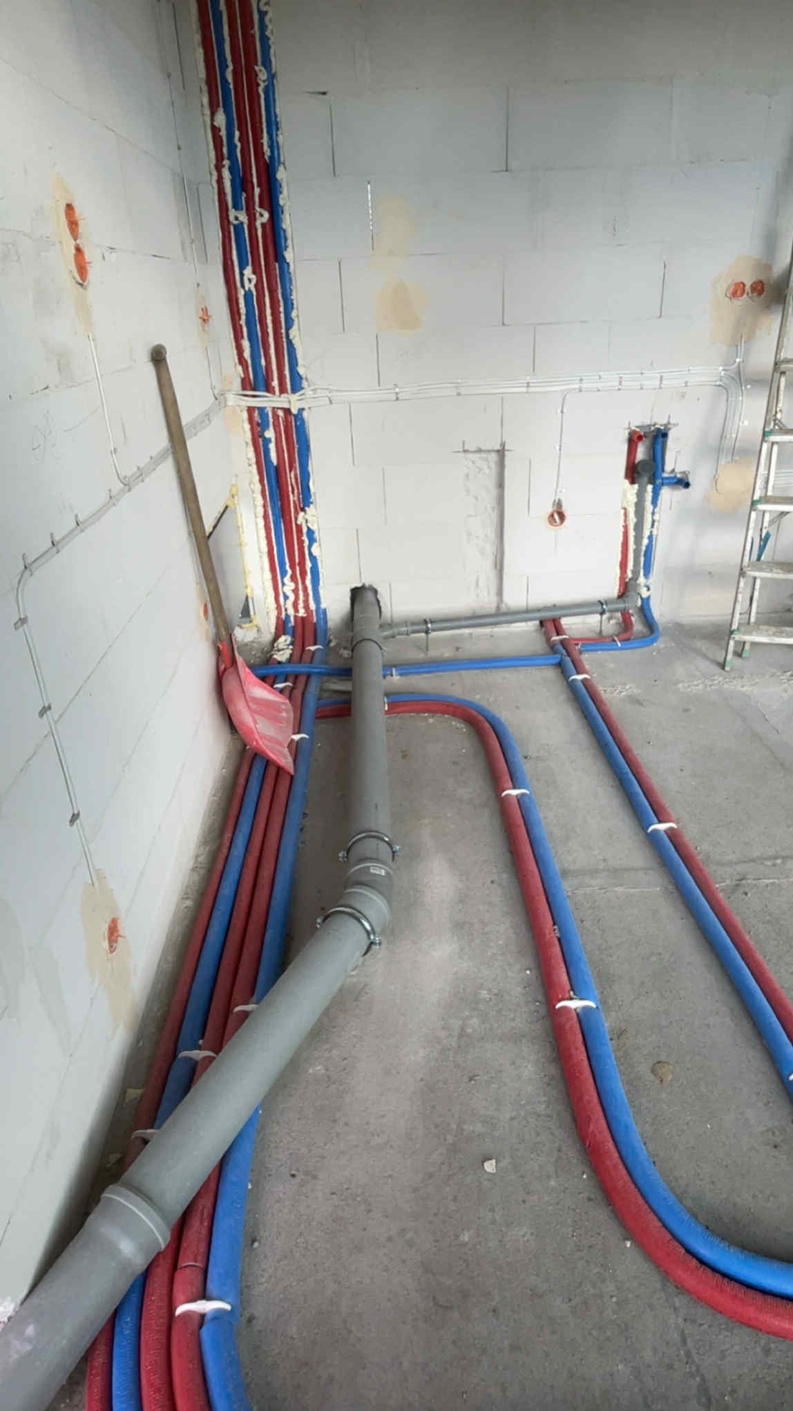 Instalacja wodna i kanalizacyjna w surowym wnętrzu: rury czerwone i niebieskie prowadzone wzdłuż ściany, szare rury kanalizacyjne na podłodze, pianka montażowa.