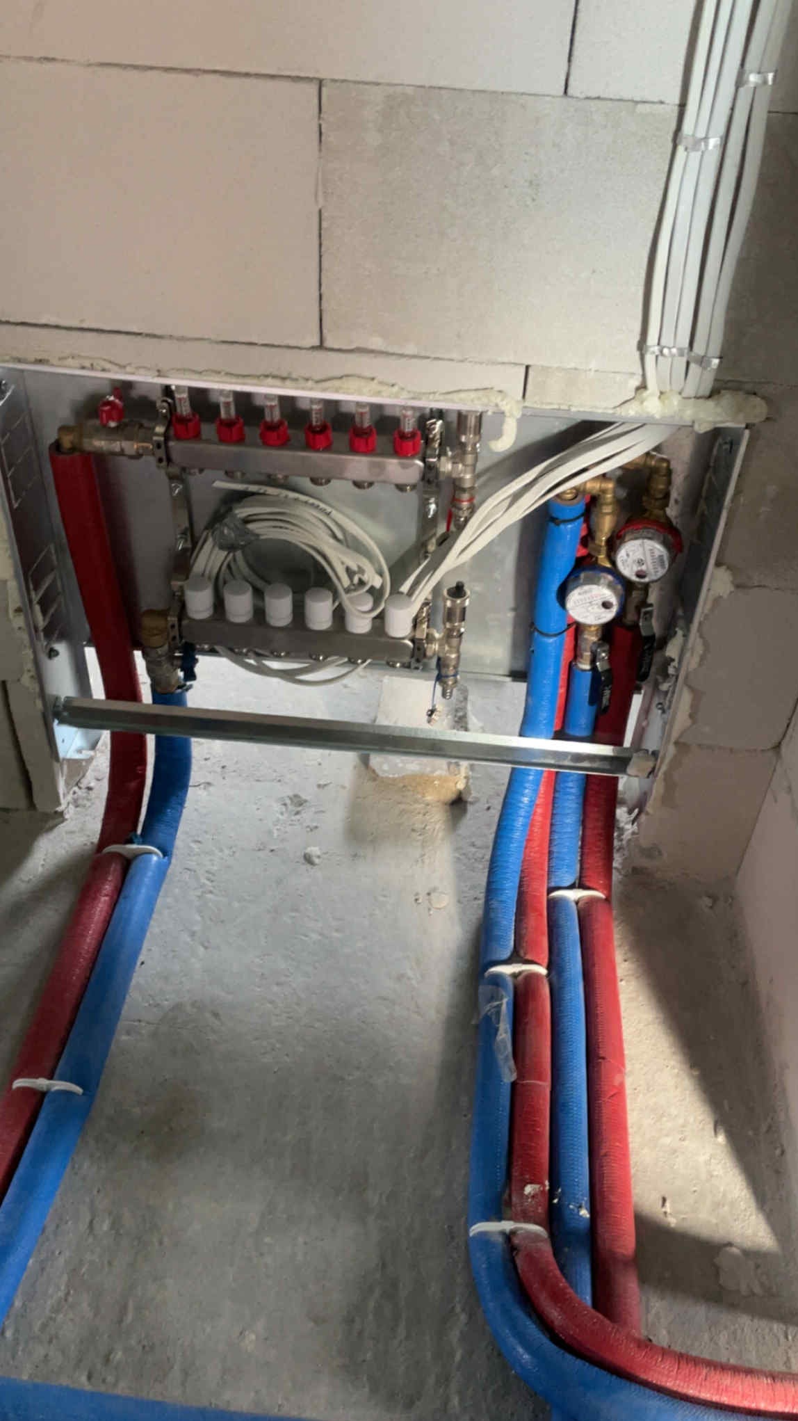 Rozdzielacz ogrzewania podłogowego z rurami w kolorze niebieskim i czerwonym, zamontowany w surowym pomieszczeniu. Widoczne białe przewody i uszczelnienia.