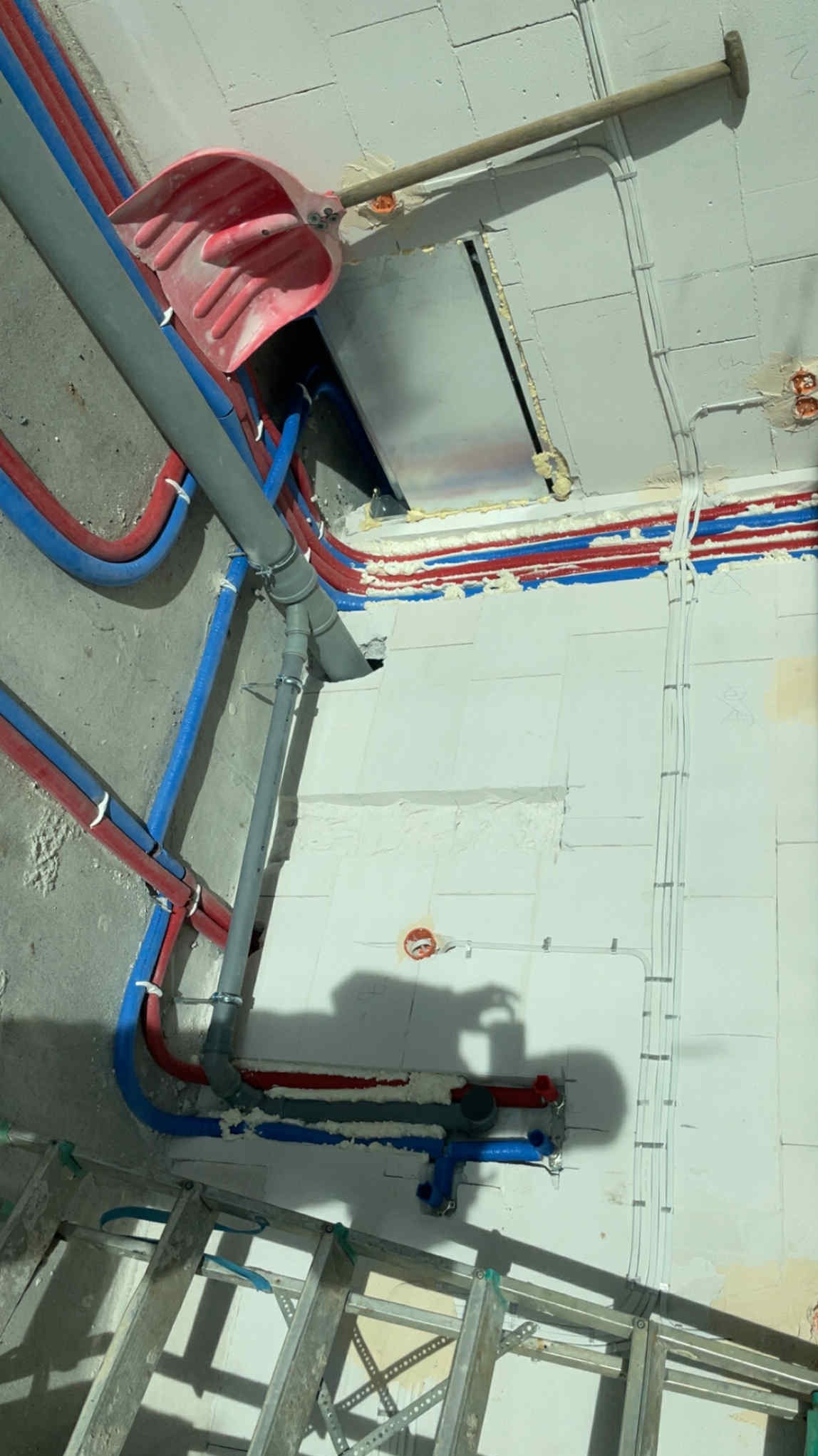 Instalacja wodna w trakcie montażu: rury w izolacji czerwono-niebieskiej, szara rura odpływowa, drabina, ściana z pustaków z instalacją elektryczną i łopatą opartą o ścianę.