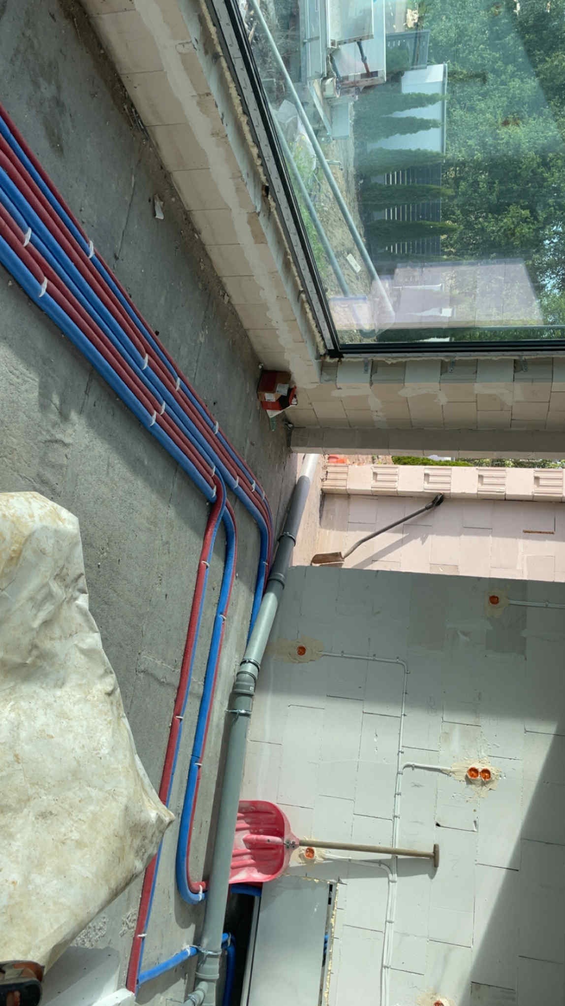 Instalacja wodna: rury czerwone i niebieskie prowadzone wzdłuż ściany z pustaków, szara rura kanalizacyjna, widoczne okablowanie elektryczne. Budowa w toku.