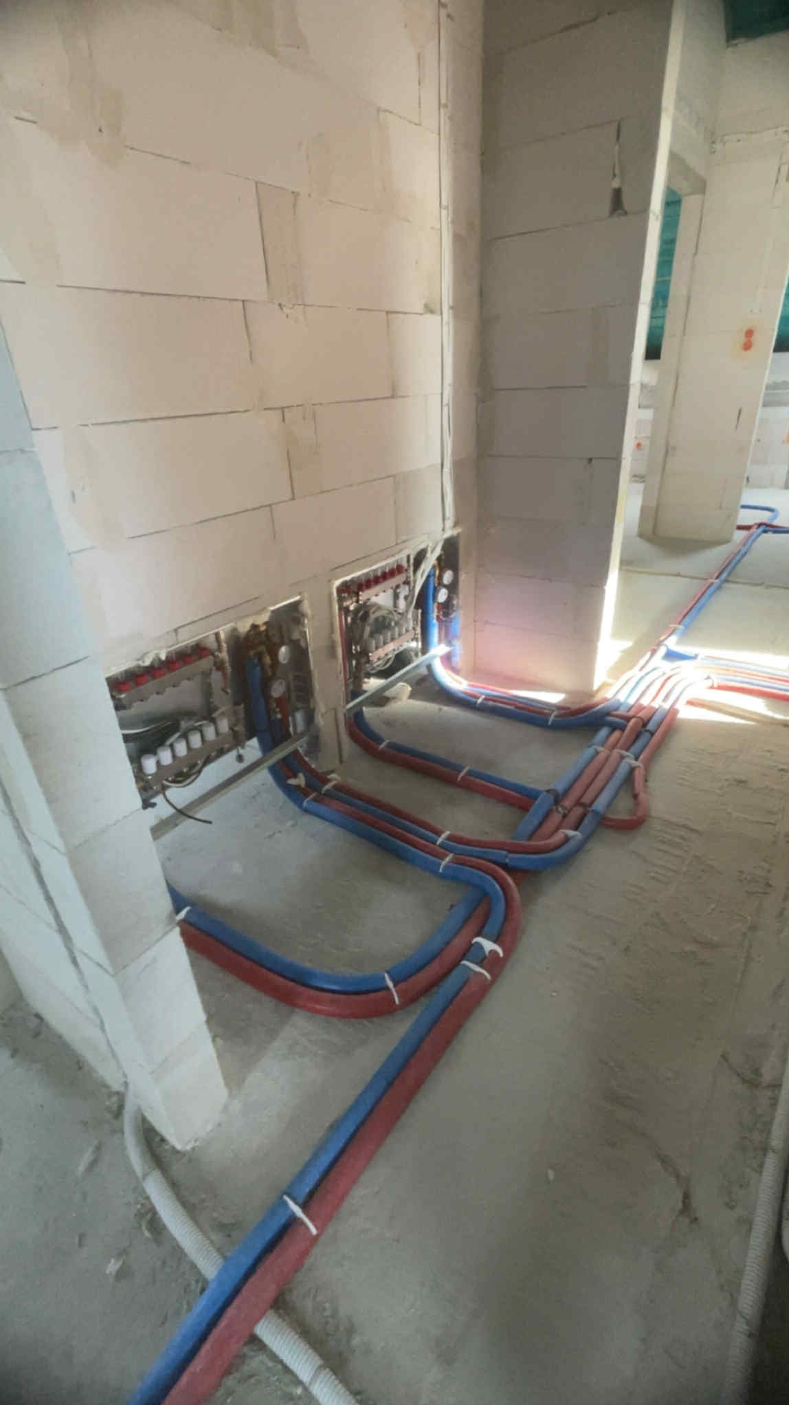 Instalacja ogrzewania podłogowego w surowym wnętrzu: czerwone i niebieskie rury prowadzone wzdłuż ścian z betonu komórkowego, widoczne rozdzielacze.