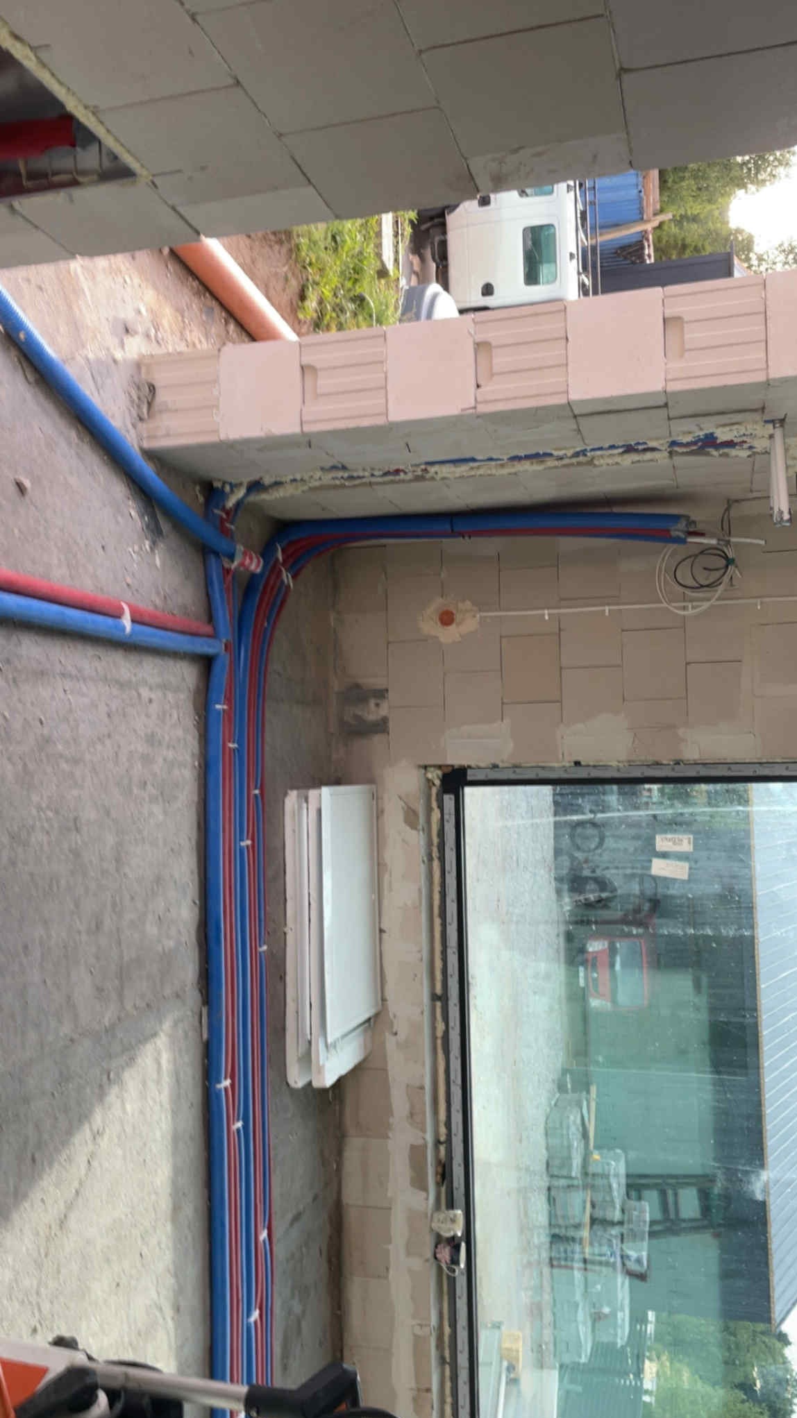 Wewnętrzne ułożenie rur instalacji wodnej i grzewczej w surowym budynku z widocznym oknem i materiałami budowlanymi na zewnątrz.