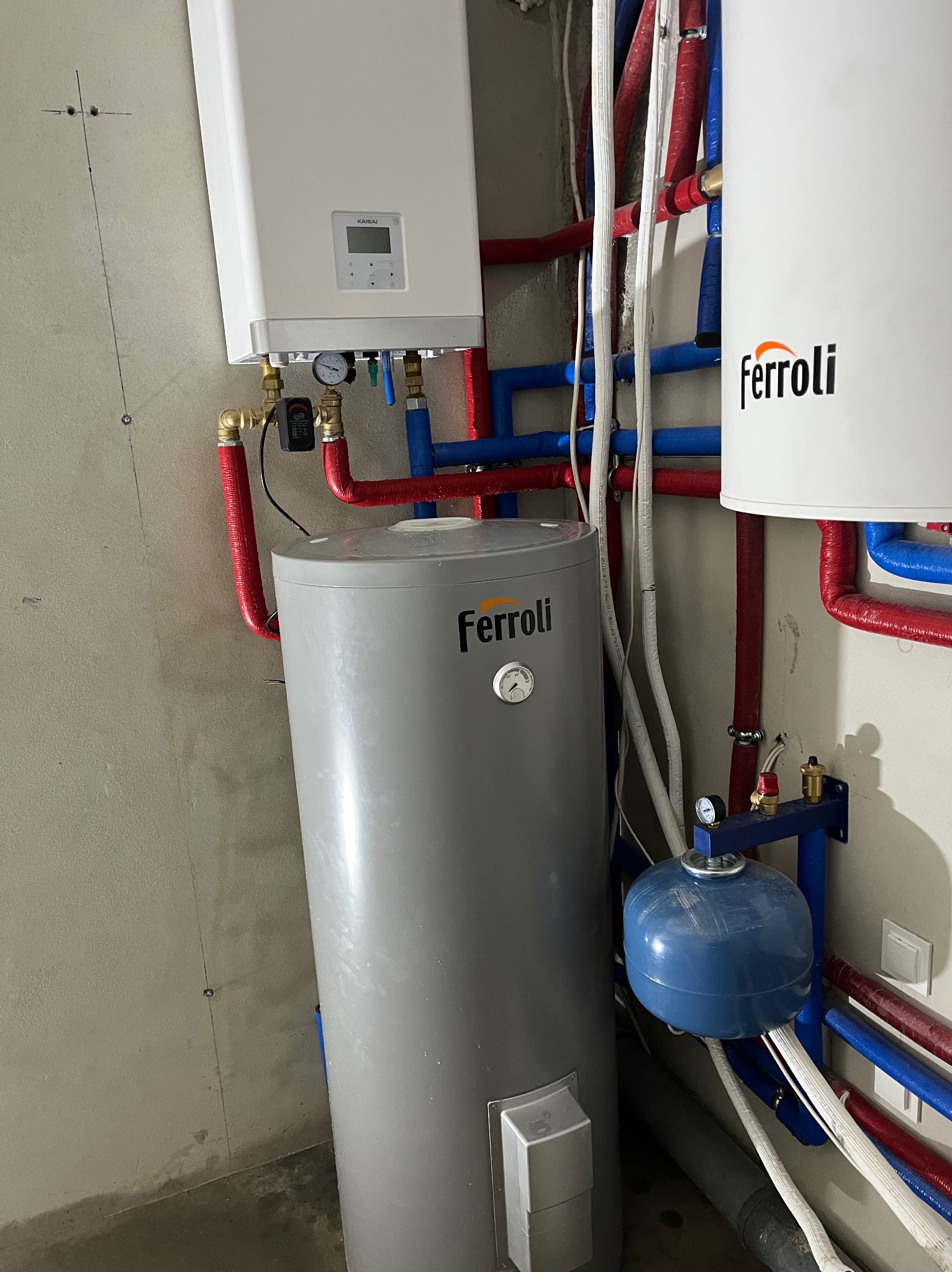 Nowoczesna instalacja grzewcza z piecem gazowym Kaisai i zasobnikami Ferroli, z widocznymi rurami w kolorach czerwonym i niebieskim, zamontowana na surowej ścianie.