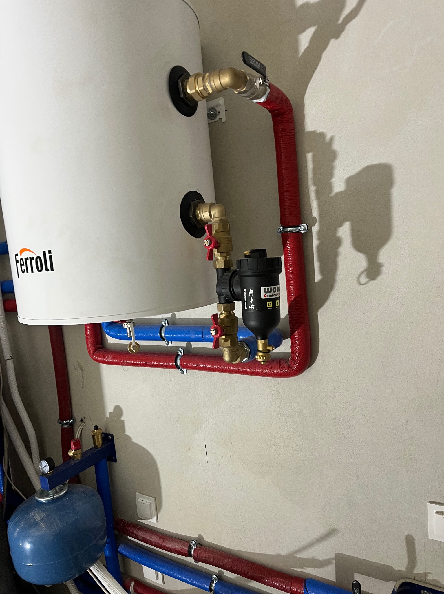 Biały zasobnik ciepłej wody Ferroli z podłączonymi rurami w izolacji czerwonej i niebieskiej, zaworami i filtrem, zamontowany na jasnej ścianie.