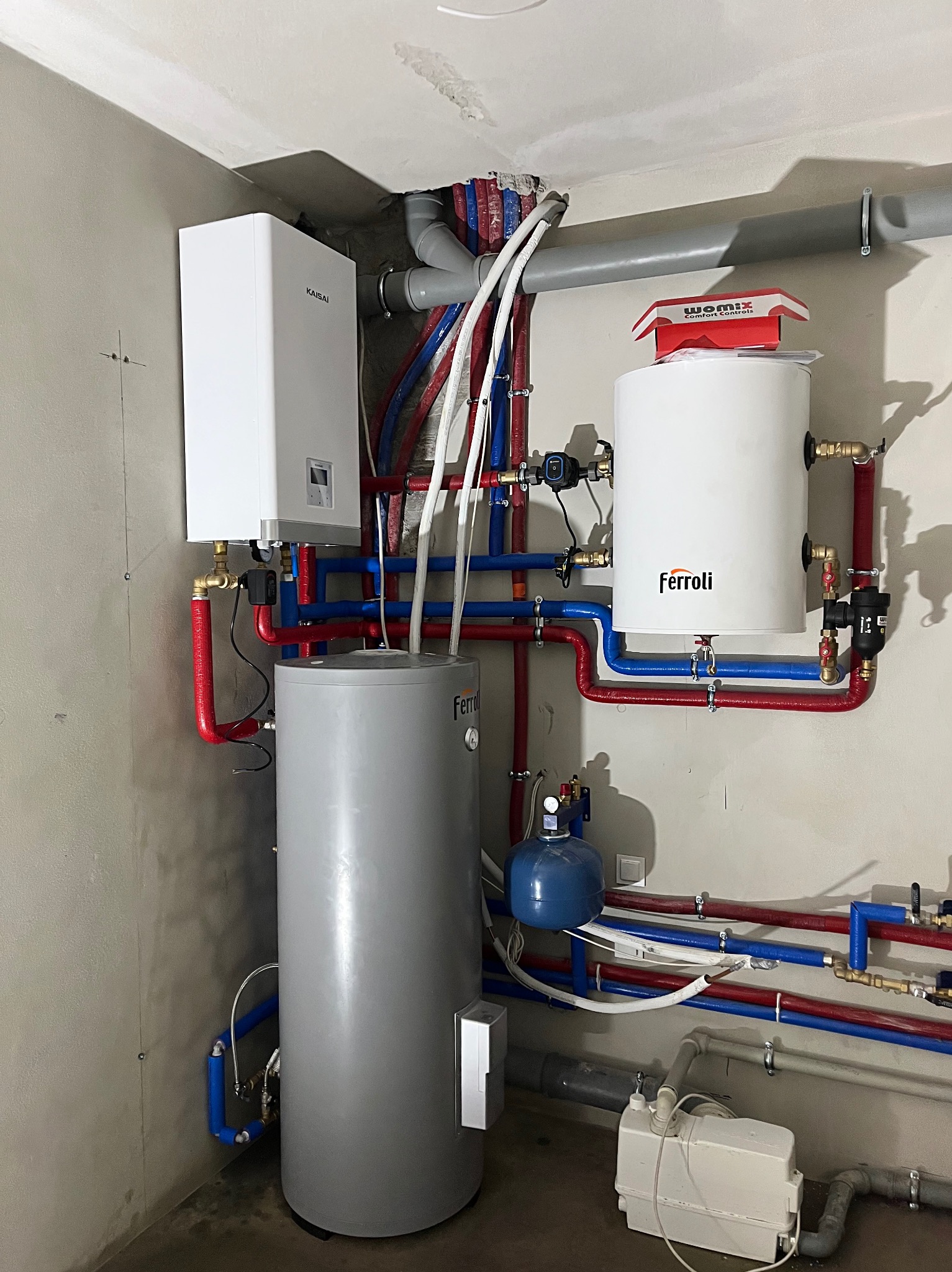 Nowoczesna kotłownia z piecem gazowym Kaisai, zasobnikiem Ferroli i instalacją z rur w kolorach czerwonym i niebieskim. Widoczne elementy armatury i osprzętu.