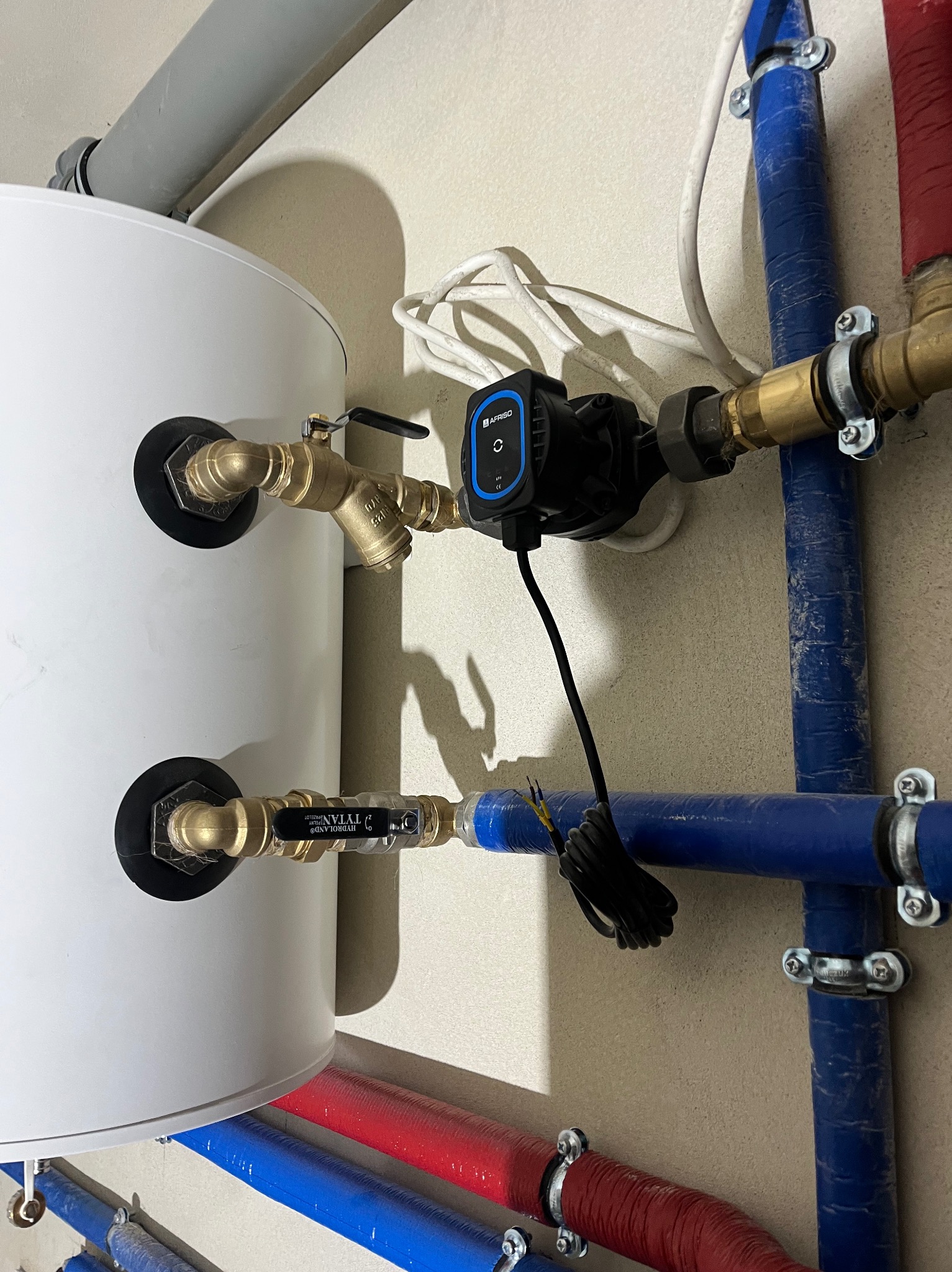 Biały bojler z podłączonymi rurami (czerwone, niebieskie) i zaworami. Widoczna pompa obiegowa Afriso. Instalacja hydrauliczna w pomieszczeniu.