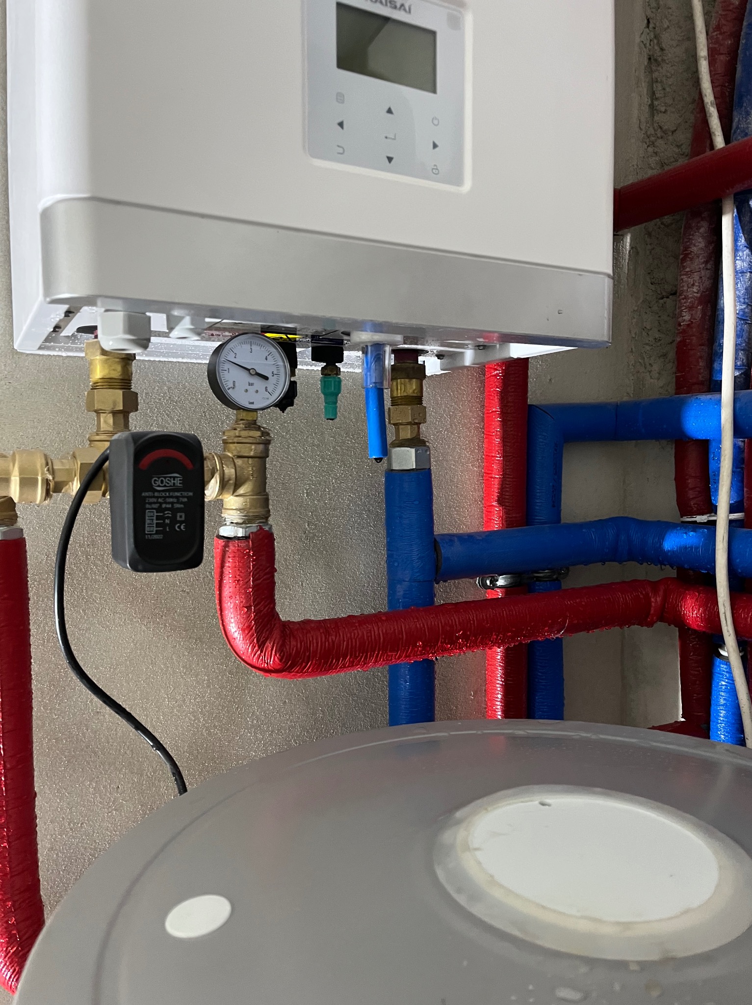 Nowoczesny piec gazowy z manometrem i zabezpieczeniem Goshe, podłączony do instalacji z rur w izolacji czerwonej i niebieskiej. Widok z dołu, detale przyłączeń.