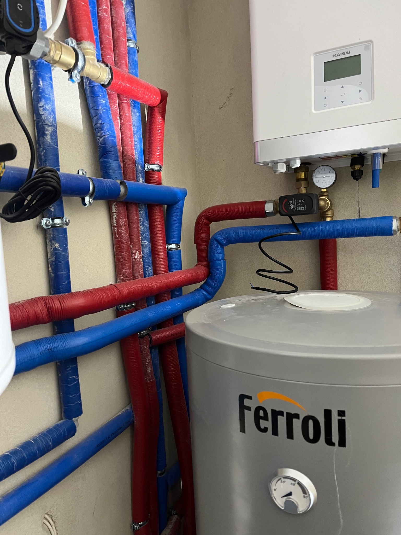 Instalacja CO z piecem Kaisai i zasobnikiem Ferroli. Rury w izolacji czerwonej i niebieskiej, zawory i czujniki. Widok na ścianę z elementami instalacji.