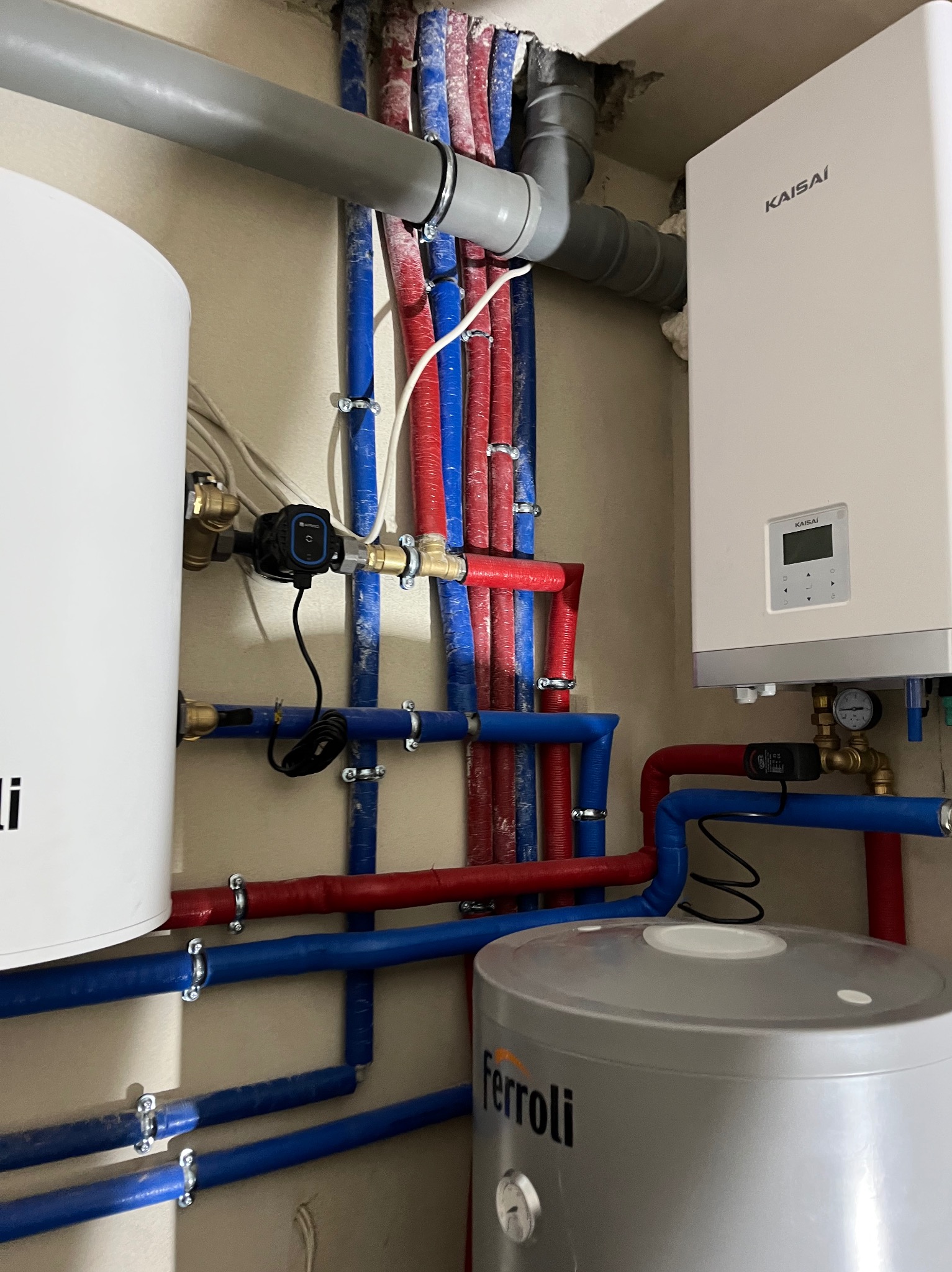 Nowoczesna kotłownia z piecem gazowym Kaisai, zasobnikiem Ferroli i rozbudowaną instalacją z rur w kolorach czerwonym i niebieskim, zamontowaną przy beżowej ścianie.