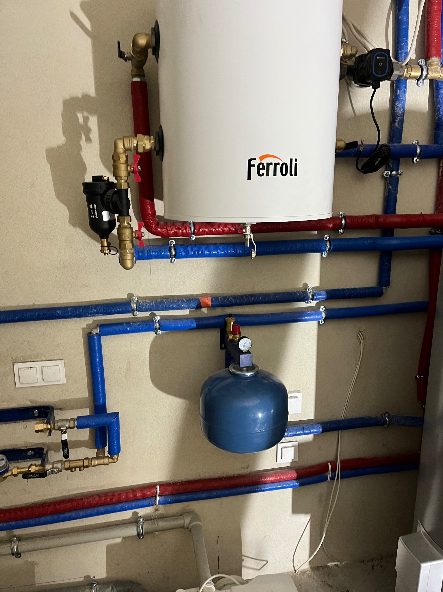 Biały piec gazowy Ferroli z podłączonymi rurami (czerwone, niebieskie) i osprzętem na tle beżowej ściany. Widoczny manometr i zawory.