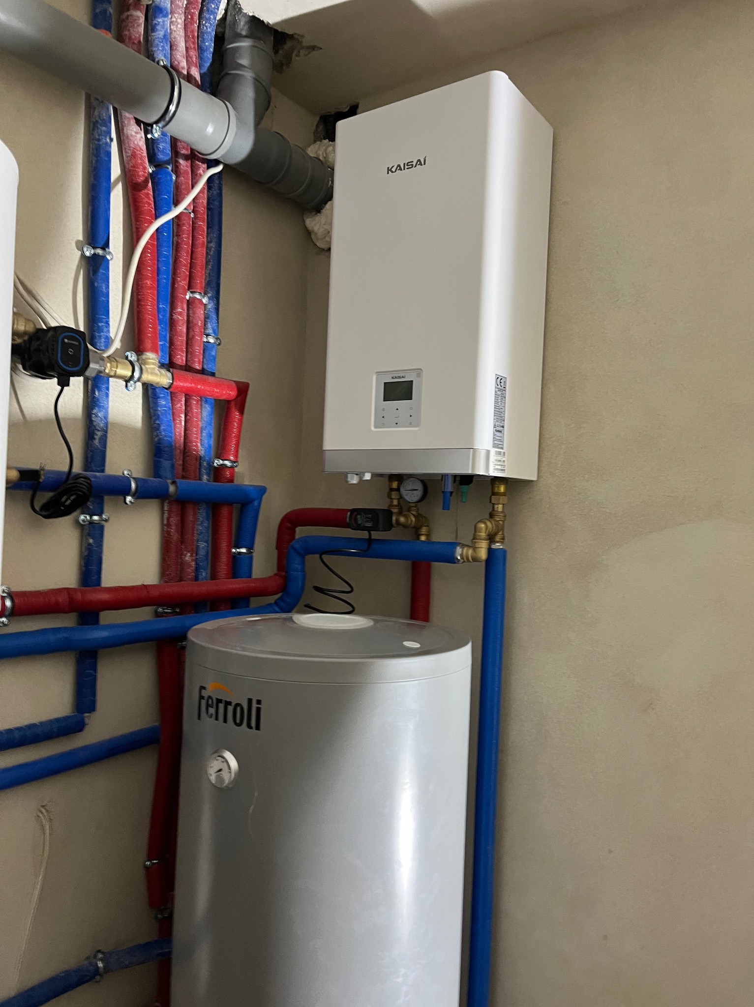 Biały piec gazowy Kaisai z szarym zasobnikiem Ferroli, połączony rurami w kolorach niebieskim i czerwonym. Instalacja w pomieszczeniu o beżowych ścianach.
