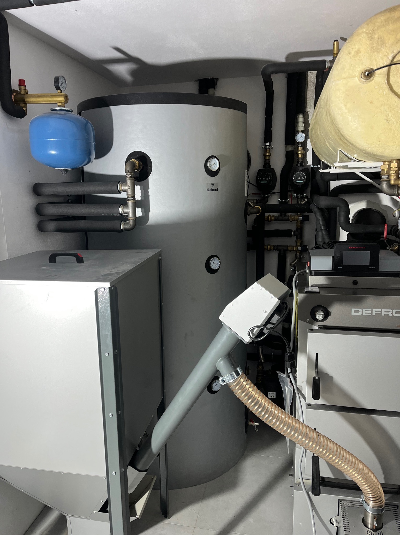 Kotłownia z piecem na paliwo stałe, zasobnikiem ciepłej wody i elementami instalacji. Widoczne rury, zawory i manometry. Całość w jasnej kolorystyce.