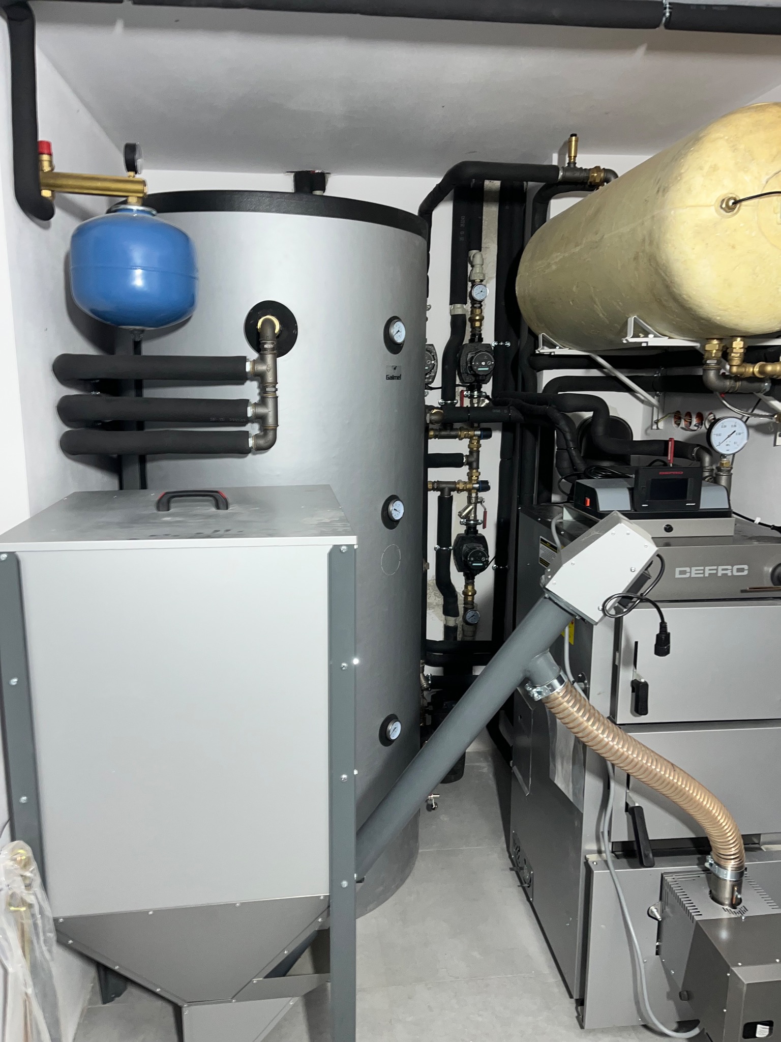 Nowoczesna kotłownia z piecem Defro, zasobnikiem i zbiornikiem wyrównawczym. Widoczne rury, zawory i manometry. Całość w jasnej, minimalistycznej aranżacji.