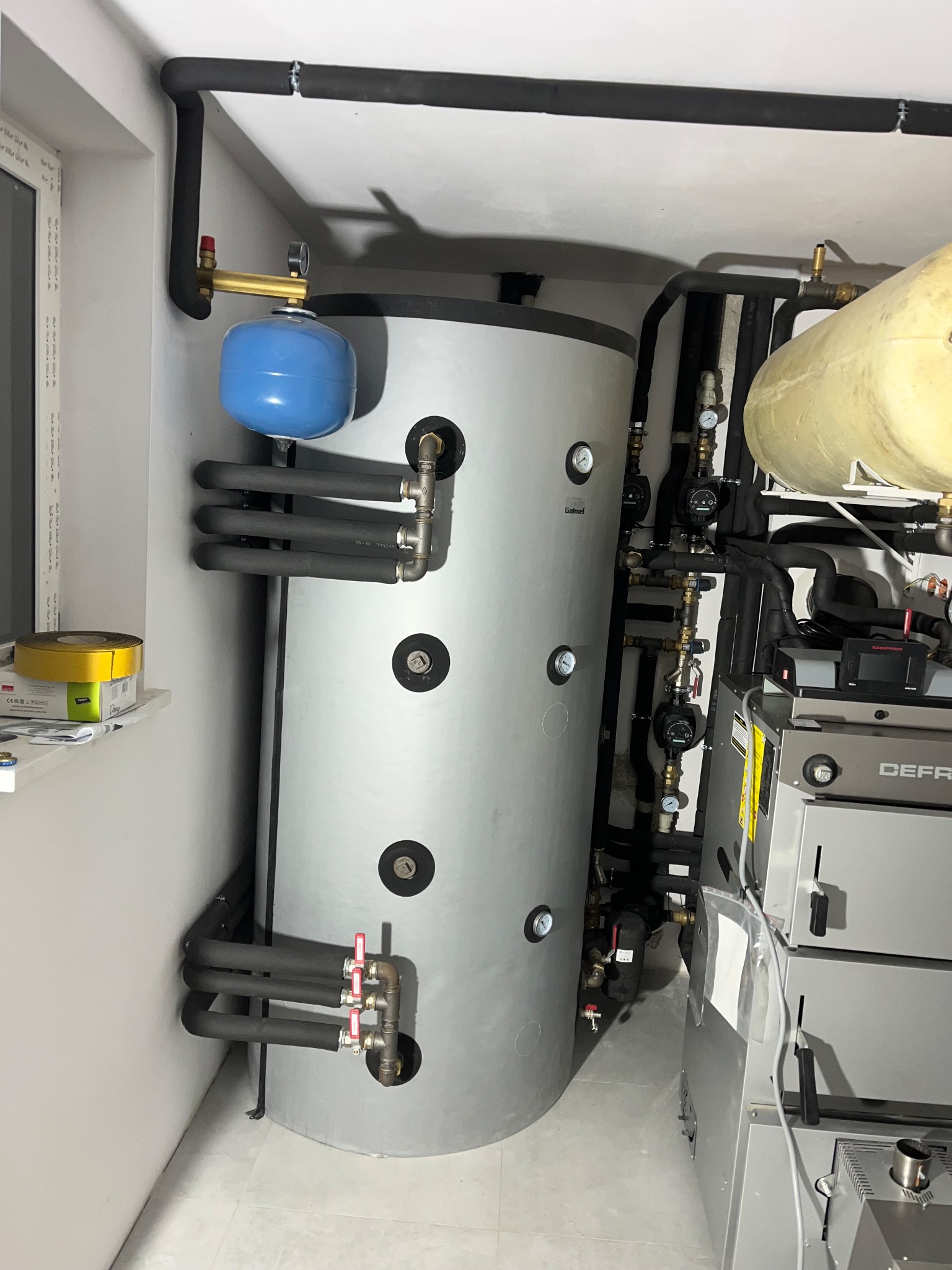 Szary, pionowy zbiornik z niebieskim naczyniem wzbiorczym i czarnymi rurami w kotłowni. Widoczne manometry i zawory. Ujęcie z bliska, oświetlenie naturalne.