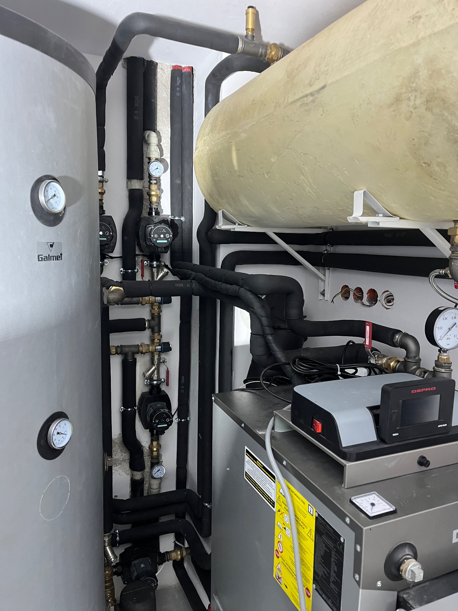 Kotłownia z piecem Defro, zbiornikiem Galmet, licznymi rurami i manometrami. Widoczne czarne izolacje termiczne na rurach. Instalacja w pomieszczeniu technicznym.